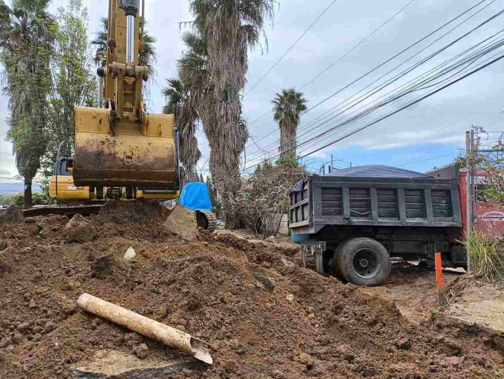 Maquinaria pesada y camión de volteo trabajando en una obra de construcción con palmeras al fondo en un área urbana